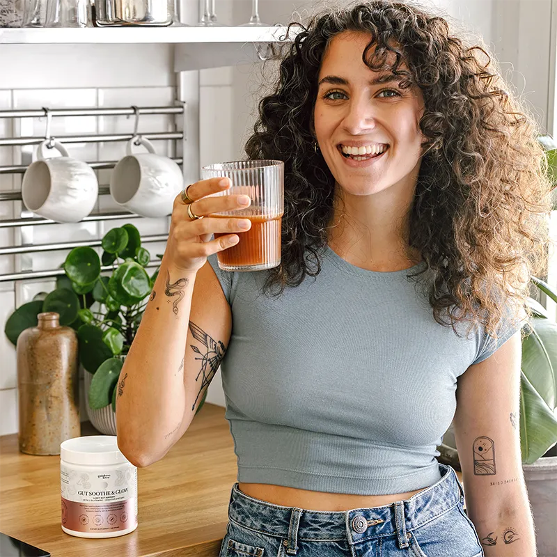 Woman enjoying a Gut Soothe & Glow smoothie in her kitchen, with the product jar on the counter beside her